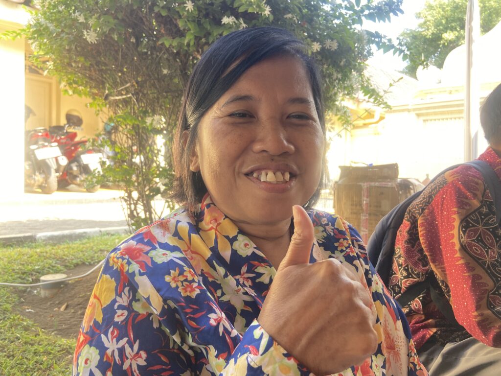 Close-up of a smiling woman in a bright floral shirt giving a thumbs-up outdoors in sunlight.