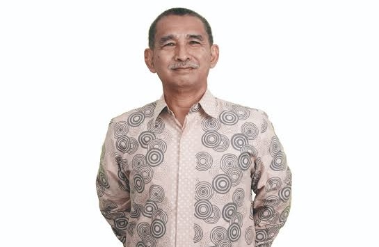 Portrait of a man in a patterned shirt standing with his hands behind his back, smiling gently.