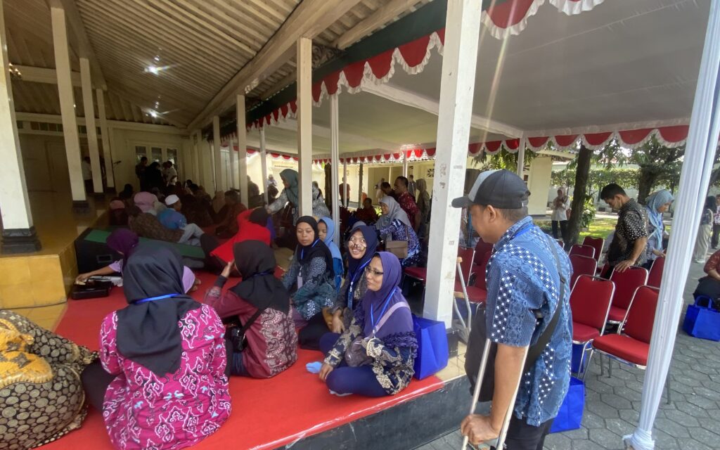 Crowd of people seated and standing under a covered walkway with red carpet and red chairs, many wearing batik and headscarves at an event.