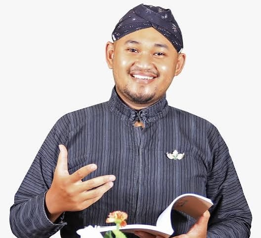Smiling man in a dark striped kurta and bandana, holding an open notebook and gesturing with his left hand.