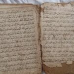 Open old book with handwritten script in Arabic on yellowed, torn pages with frayed edges.