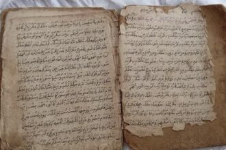 Open old book with handwritten script in Arabic on yellowed, torn pages with frayed edges.