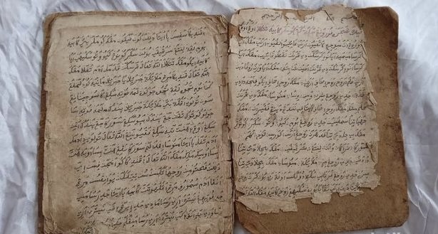 Open old book with handwritten script in Arabic on yellowed, torn pages with frayed edges.
