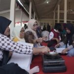 Group of women in hijabs sit and stand around a small gas stove cooking on a red carpet inside a tented hall.