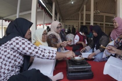 Group of women in hijabs sit and stand around a small gas stove cooking on a red carpet inside a tented hall.