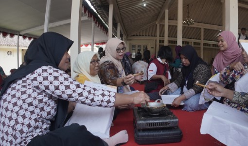 Group of women in hijabs sit and stand around a small gas stove cooking on a red carpet inside a tented hall.