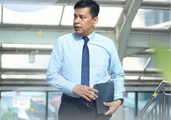 IMG_3932 - Mabur.co Man in a light blue shirt and navy tie standing with a folder in a bright, modern office lobby with glass walls.