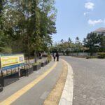 Wide daytime street scene with a curved pedestrian path, signboard on the left, trees, and a building in the distance in blue sky.