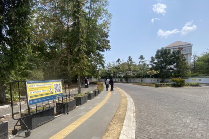 Wide daytime street scene with a curved pedestrian path, signboard on the left, trees, and a building in the distance in blue sky.