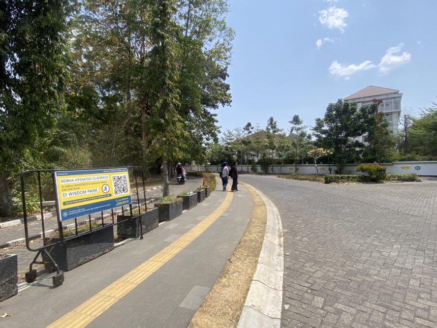 Wide daytime street scene with a curved pedestrian path, signboard on the left, trees, and a building in the distance in blue sky.