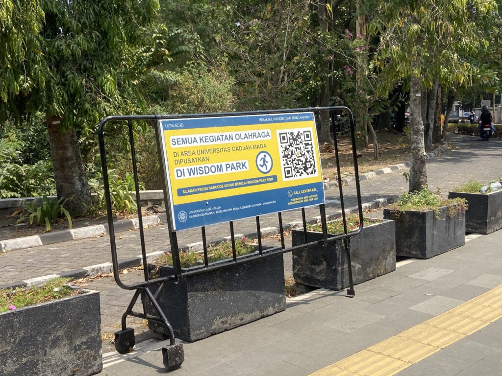 Yellow and blue signboard on a metal frame beside a tree-lined walkway, advertising sports activities at Wisdom Park. близ