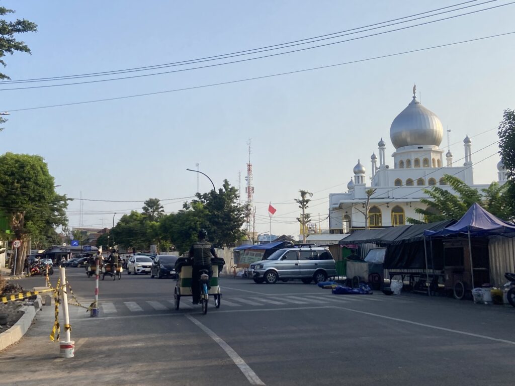 IMG_3945 - Mabur.co Street view with a white mosque featuring a large silver dome on the right and minarets; traffic includes cars, motorbikes, and a rickshaw in the foreground under a clear blue sky