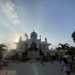 White mosque with a blue central dome and crescent finial, sun flares in the foreground, and visitors around the entrance on a sunny day.