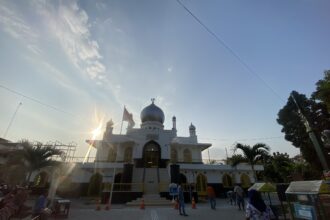 White mosque with a blue central dome and crescent finial, sun flares in the foreground, and visitors around the entrance on a sunny day.