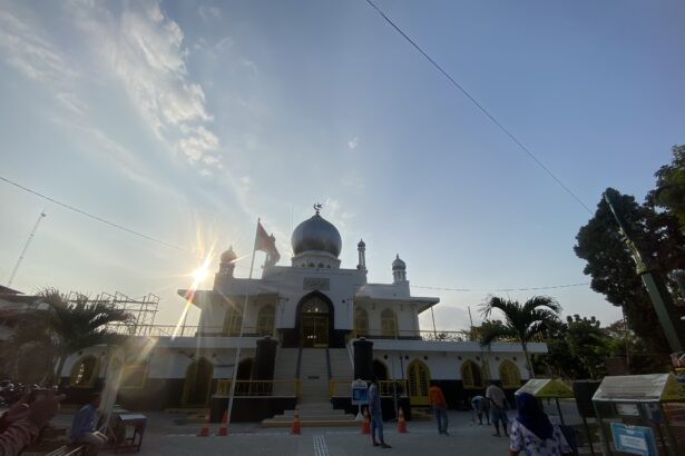 White mosque with a blue central dome and crescent finial, sun flares in the foreground, and visitors around the entrance on a sunny day.