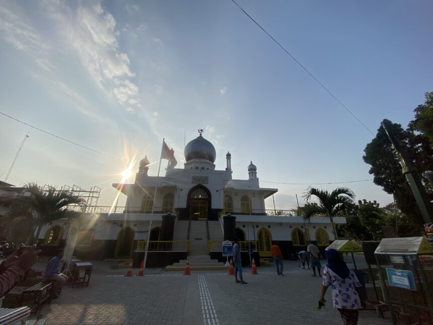 White mosque with a blue central dome and crescent finial, sun flares in the foreground, and visitors around the entrance on a sunny day.