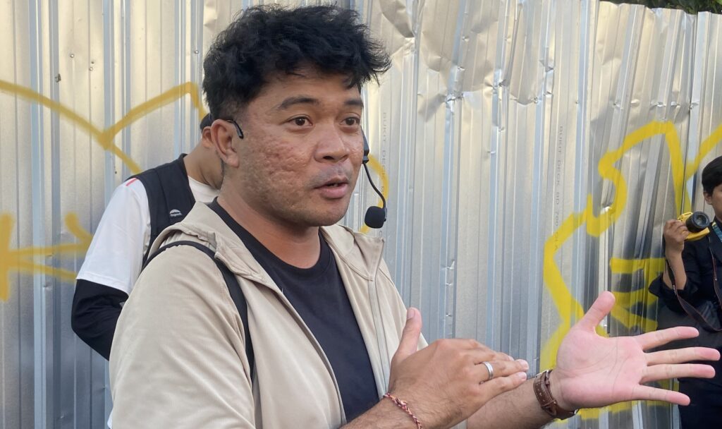 Man wearing a headset mic and beige jacket gestures with hands in front of a graffiti-covered metal wall.