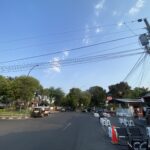 Urban street scene with utility poles and a tangle of power lines against a clear blue sky; trees line the left side and a small setup of white chairs and barriers on the right.