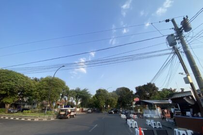 Urban street scene with utility poles and a tangle of power lines against a clear blue sky; trees line the left side and a small setup of white chairs and barriers on the right.