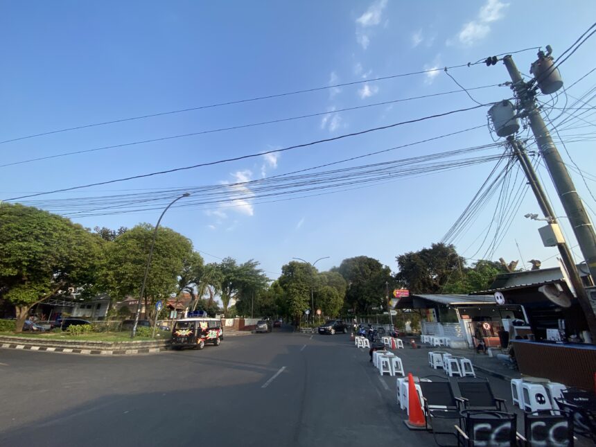 Urban street scene with utility poles and a tangle of power lines against a clear blue sky; trees line the left side and a small setup of white chairs and barriers on the right.