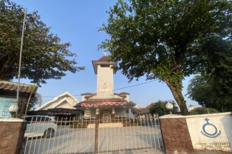 Front view of a white church tower with a cross, framed by green trees and a metal gate.
