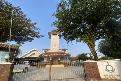 Front view of a white church tower with a cross, framed by green trees and a metal gate.