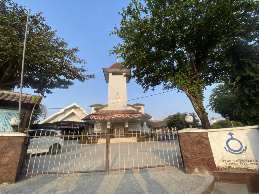 Front view of a white church tower with a cross, framed by green trees and a metal gate.