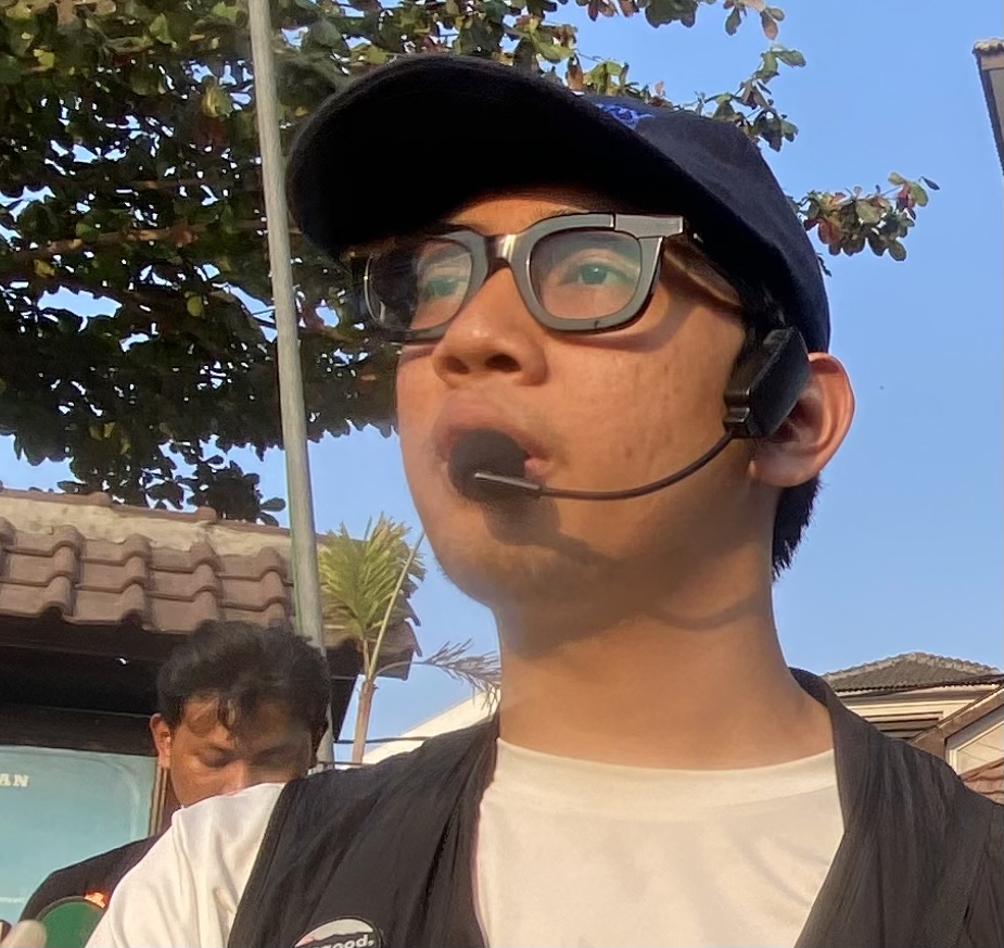 IMG_3960 - Mabur.co Close-up of a young man wearing a black cap, glasses, and a headset microphone, speaking outdoors in daylight.
