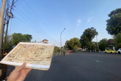 Person holding an open map/brochure over a dark street with trees, power lines, and a few cars in the background