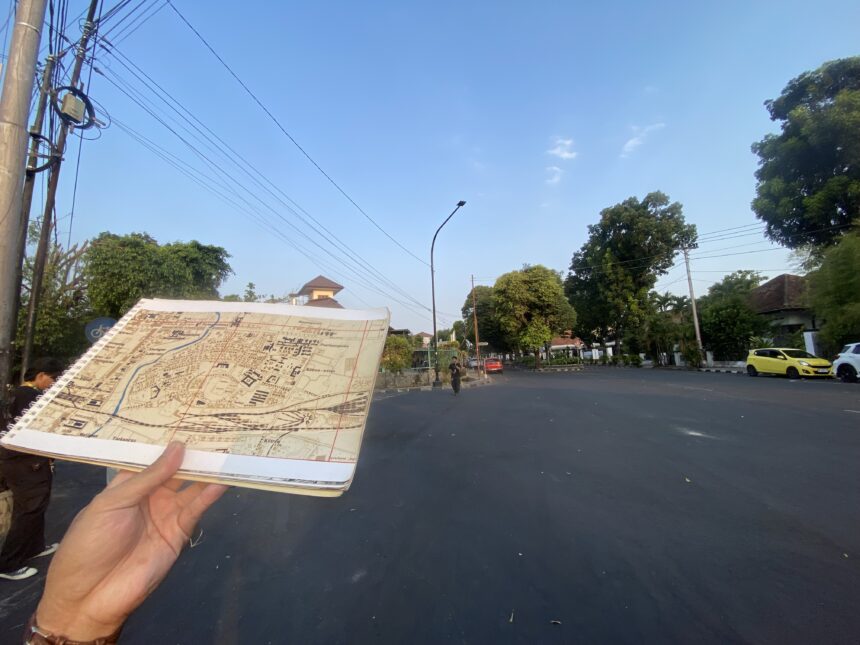 Person holding an open map/brochure over a dark street with trees, power lines, and a few cars in the background