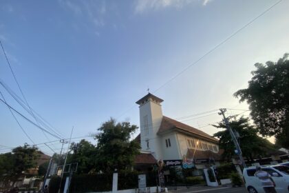 Urban street view with a white building and red-tiled roof, power lines crossing a clear blue sky, trees along the sidewalk.
