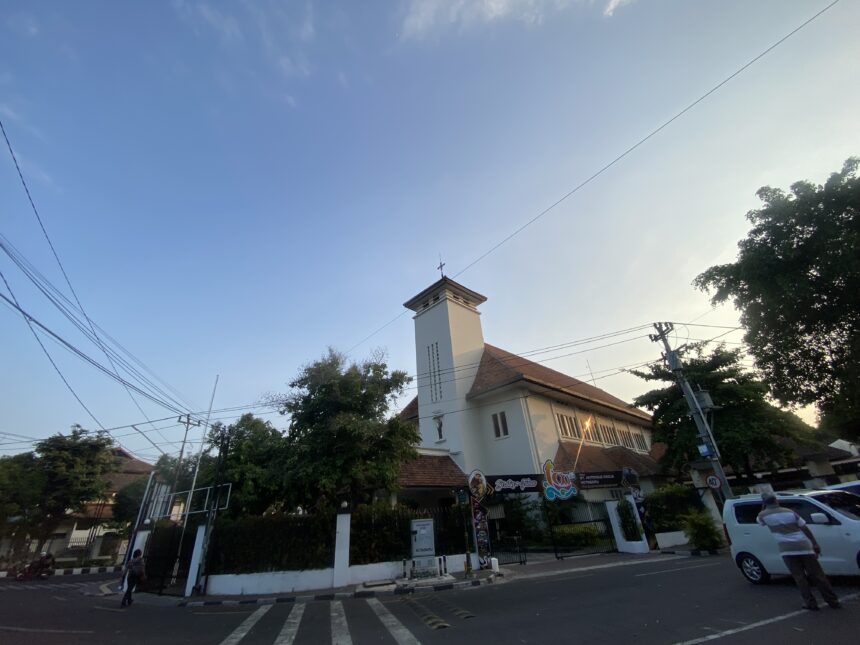 Urban street view with a white building and red-tiled roof, power lines crossing a clear blue sky, trees along the sidewalk.