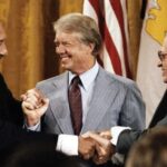 Three men in business suits smiling and clasping hands in a group handshake at a formal event, with flags in the background.