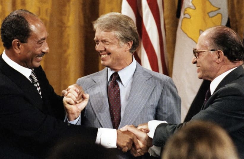Three men in business suits smiling and clasping hands in a group handshake at a formal event, with flags in the background.