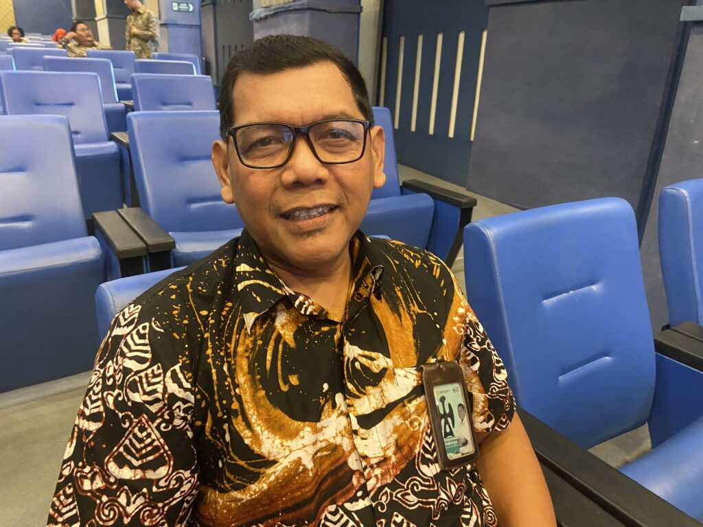 IMG_4018 - Mabur.co Self-portrait of a man wearing glasses and a batik shirt, sitting in blue theater seats inside a conference hall.