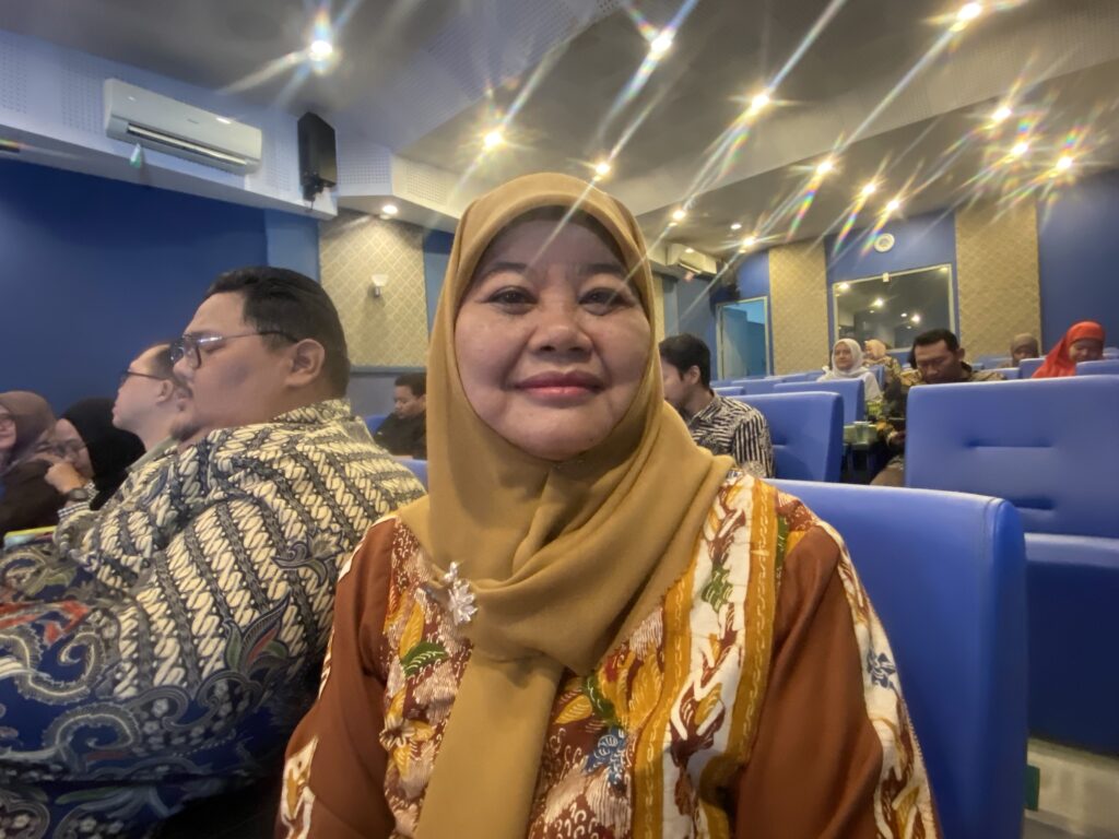 IMG_4024 - Mabur.co Woman in a mustard hijab taking a selfie in a blue, auditorium-style seating area with other passengers in patterned clothing in the background.