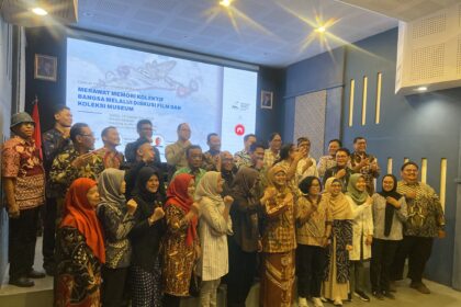Group of people in batik clothing posing on a stage or platform, smiling for a group photo with a projection screen in the background.