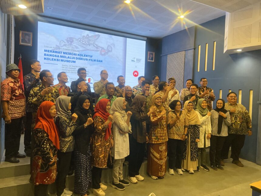 Group of people in batik clothing posing on a stage or platform, smiling for a group photo with a projection screen in the background.