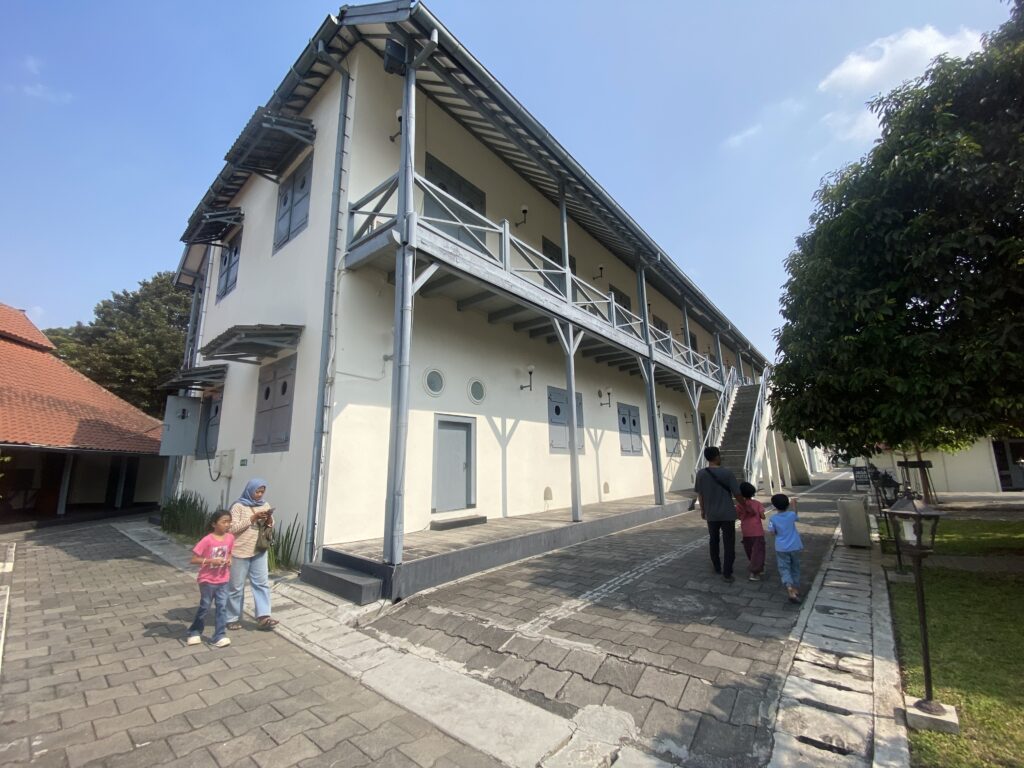 IMG_4040 - Mabur.co Three-story beige building with blue-gray balconies and a wide staircase, people walking in a paved courtyard under a clear blue sky.