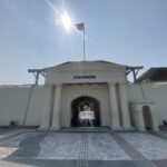 Entrance to Vredeburg fort with white columns, an arched gateway, and a flag atop a tall pole under a bright sun.