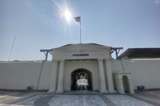Entrance to Vredeburg fort with white columns, an arched gateway, and a flag atop a tall pole under a bright sun.