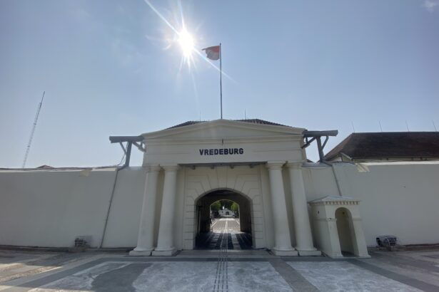 Entrance to Vredeburg fort with white columns, an arched gateway, and a flag atop a tall pole under a bright sun.