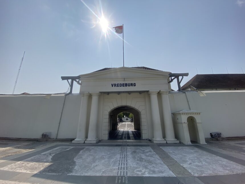 Entrance to Vredeburg fort with white columns, an arched gateway, and a flag atop a tall pole under a bright sun.