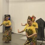 Group of young women in bright yellow traditional costumes dancing on a stage, hands raised in graceful poses with floral headpieces and colorful skirts.