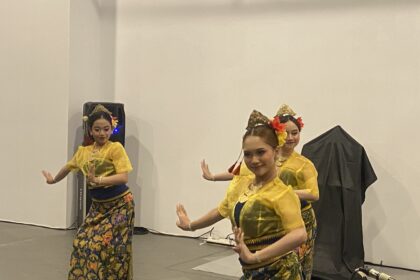 Group of young women in bright yellow traditional costumes dancing on a stage, hands raised in graceful poses with floral headpieces and colorful skirts.