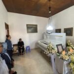 Funeral room with a white casket surrounded by white and yellow flowers; several mourners stand along the walls, facing the casket.