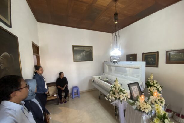 Funeral room with a white casket surrounded by white and yellow flowers; several mourners stand along the walls, facing the casket.