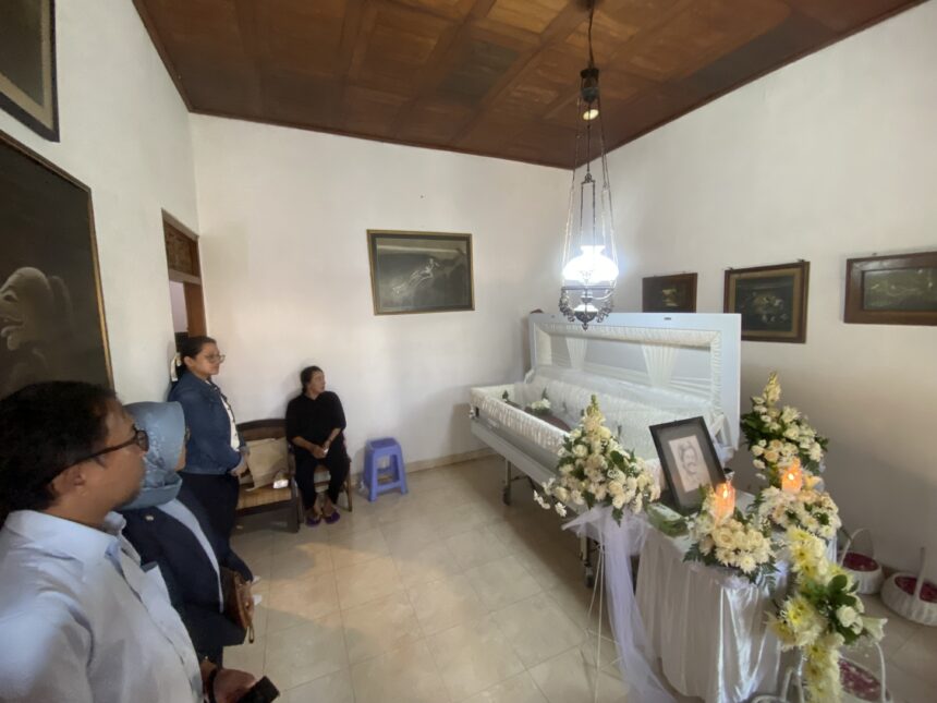 Funeral room with a white casket surrounded by white and yellow flowers; several mourners stand along the walls, facing the casket.