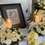 Framed portrait of a smiling person surrounded by white flowers and glowing candles at a funeral arrangements display