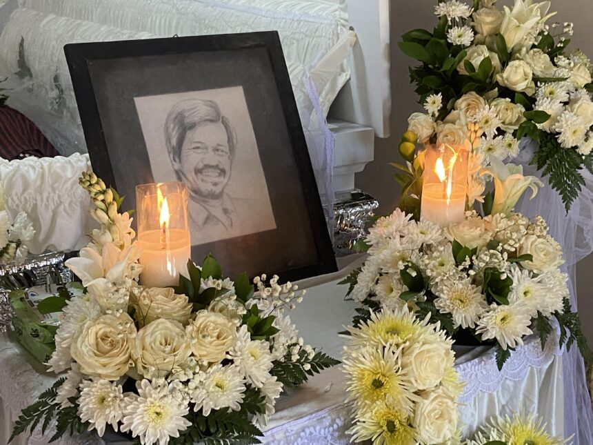 Framed portrait of a smiling person surrounded by white flowers and glowing candles at a funeral arrangements display